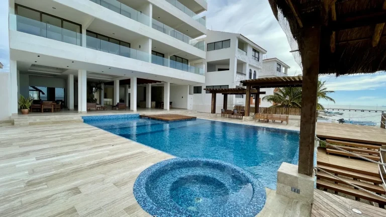 Alberca azul con sección circular junto a un edificio blanco con balcones, pérgolas y vista al mar.