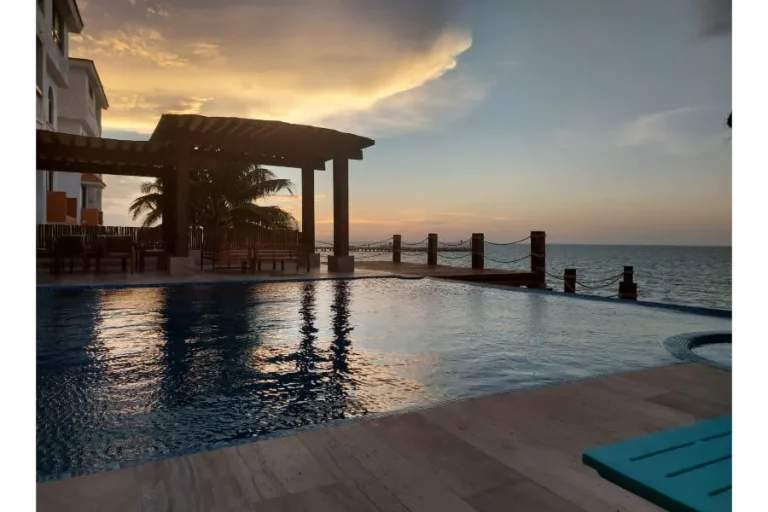 Alberca frente al mar al atardecer, con pérgola de madera, palmeras y reflejos del cielo sobre el agua.