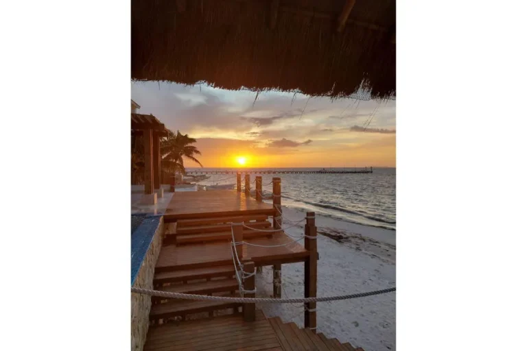Sol cerca del horizonte sobre el mar, con terrazas de madera, barandales de cuerda y playa bajo una palapa.