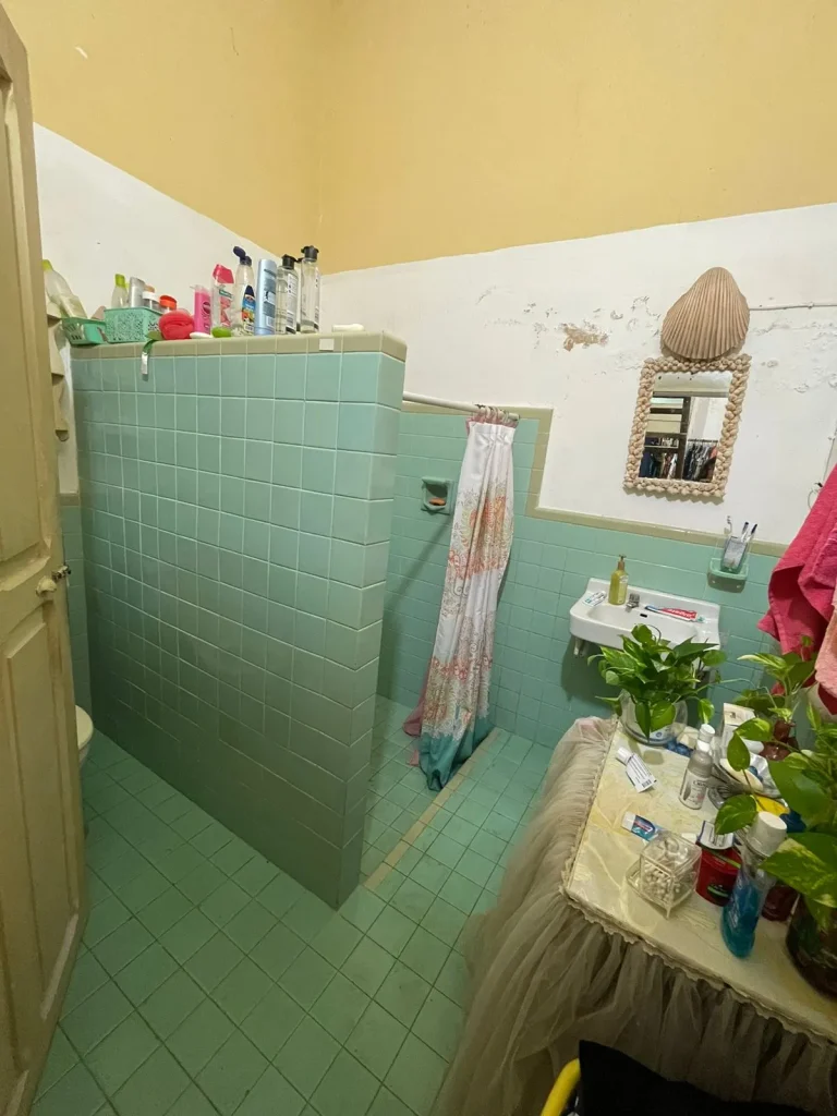 Baño con azulejos verdes, ducha con cortina, lavabo pequeño y espejo con marco de conchas.