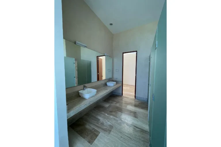 Baño con dos lavabos blancos sobre una cubierta de piedra, espejo amplio, piso de piedra y cancel de cristal.