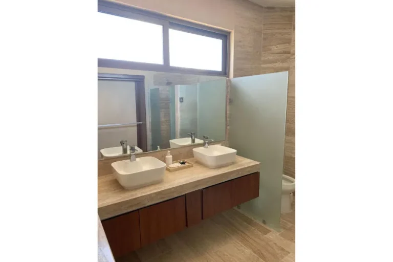 Baño con dos lavabos sobre cubierta de piedra, espejo amplio, mueble de madera y división de vidrio esmerilado.