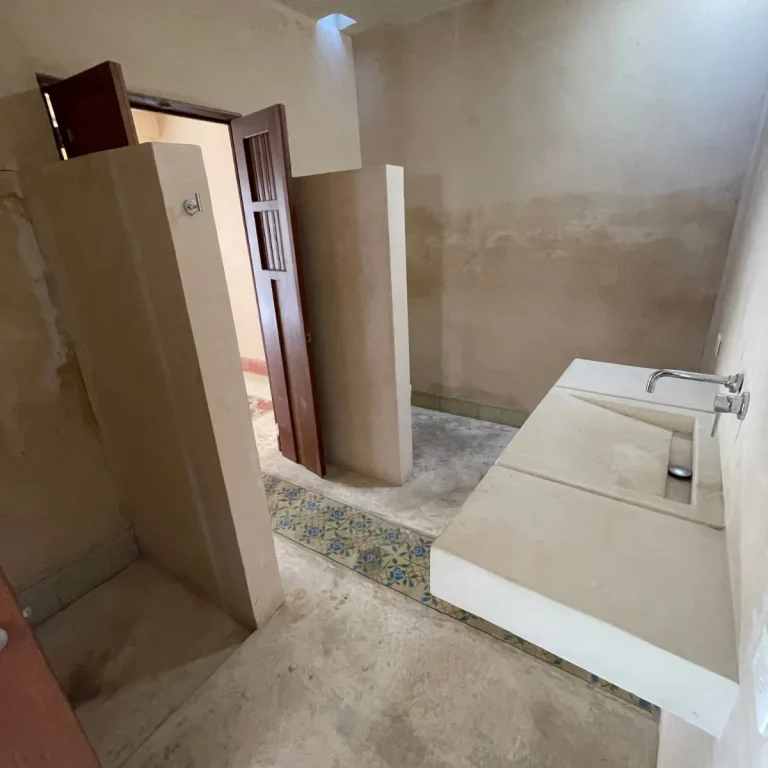Baño con lavabo rectangular integrado, grifería de pared, muros divisorios beige y una franja de mosaicos en el piso.