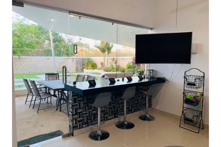 Barra negra con tres bancos, televisor en la pared y puertas de vidrio hacia una terraza con comedor y jardín.
