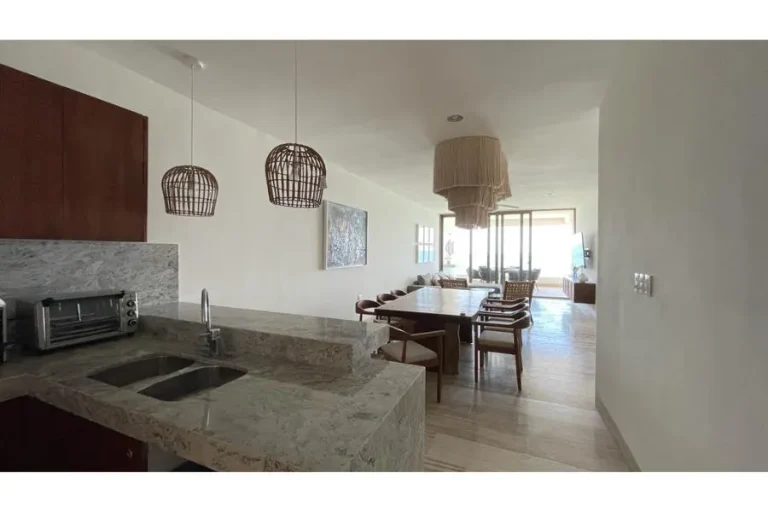 Barra de cocina con fregadero en primer plano y comedor de madera al fondo, bajo lámparas colgantes de fibras naturales.