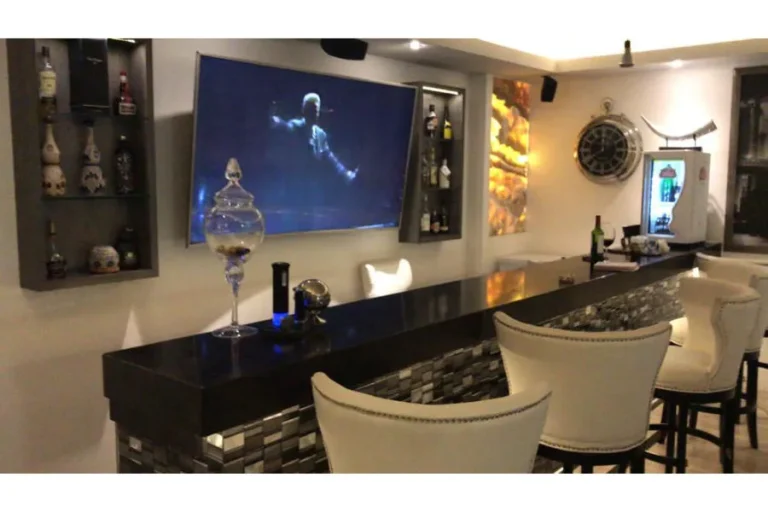 Barra negra con bancos tapizados claros, vitrinas con botellas y pantalla en la pared.