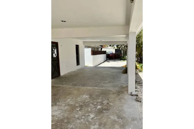 Cochera techada con columnas y paredes blancas, piso de concreto y acceso hacia un portón al fondo.