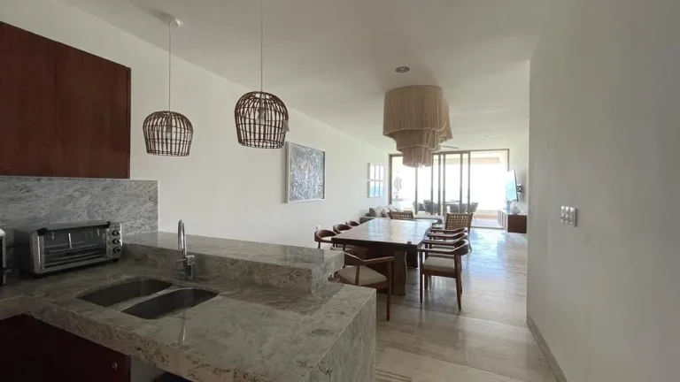 Cocina abierta con barra de piedra y fregadero, comedor de madera, lámparas colgantes y sala al fondo.