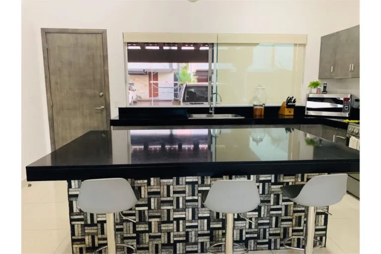 Cocina con barra negra brillante, frente de mosaico geométrico, tres bancos blancos y fregadero bajo la ventana.