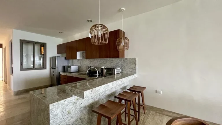 Cocina con barra de piedra gris, tres bancos de madera, gabinetes de madera y lámparas colgantes de fibras.
