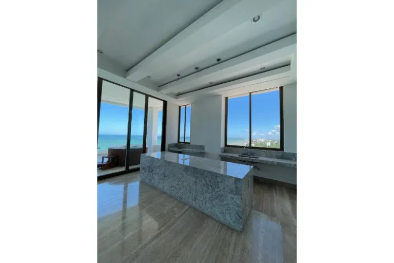 Cocina con isla de acabado pétreo, fregadero bajo la ventana y vista al mar a través de puertas de cristal.