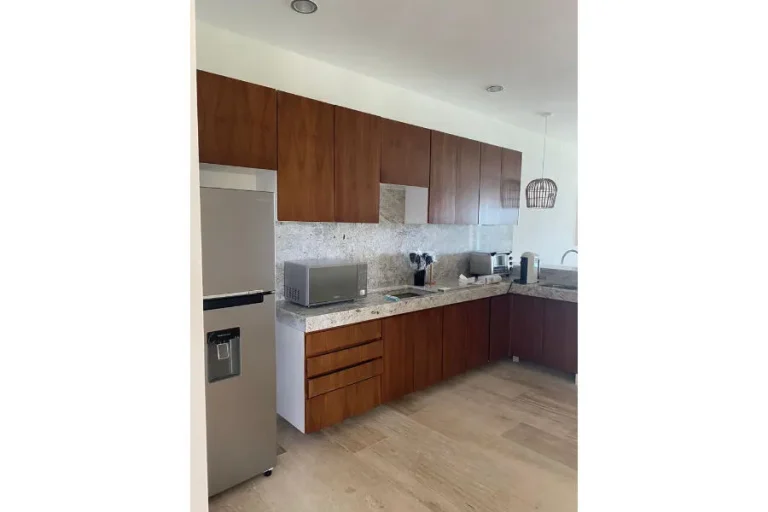 Cocina con gabinetes de madera, cubierta clara, refrigerador, microondas y lámpara colgante sobre el fregadero.