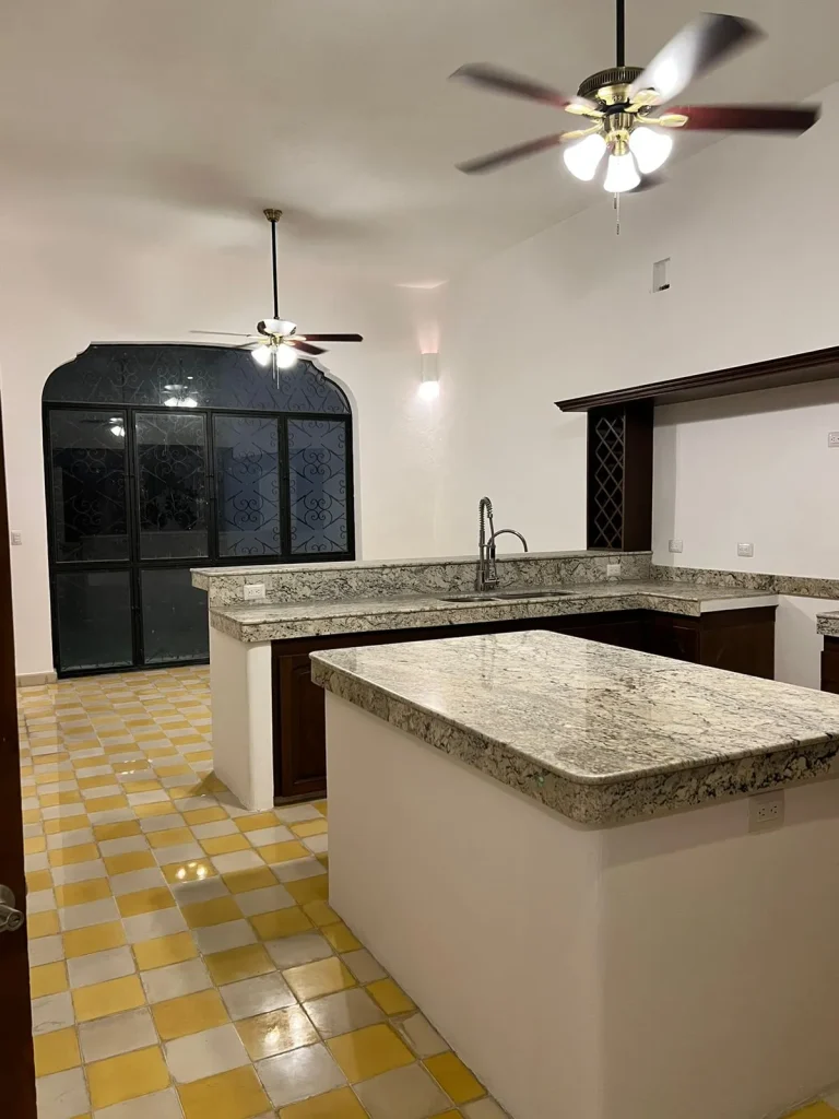 Cocina con isla, cubiertas de piedra, gabinetes oscuros y piso de cuadros amarillos y blancos.