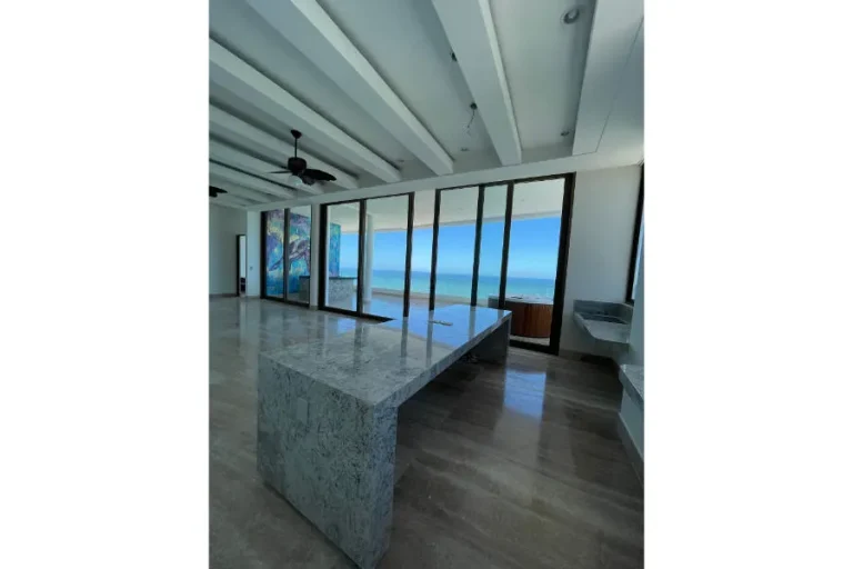Cocina con amplia isla de piedra, vigas blancas y ventanales de piso a techo con vista al mar.