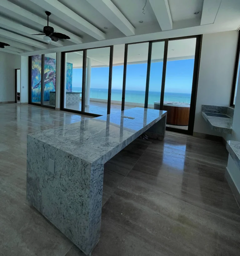Cocina con isla de piedra veteada, ventanales hacia una terraza y vista al mar.