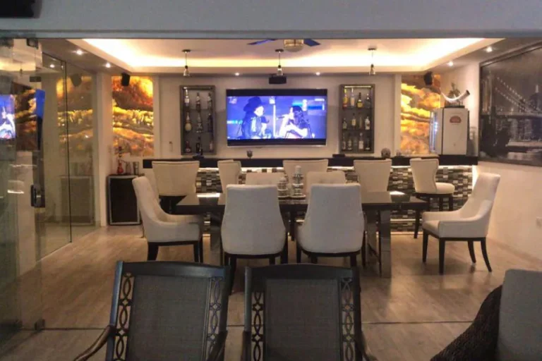 Comedor con mesa y sillas tapizadas claras, barra al fondo, televisión central y vitrinas con botellas.