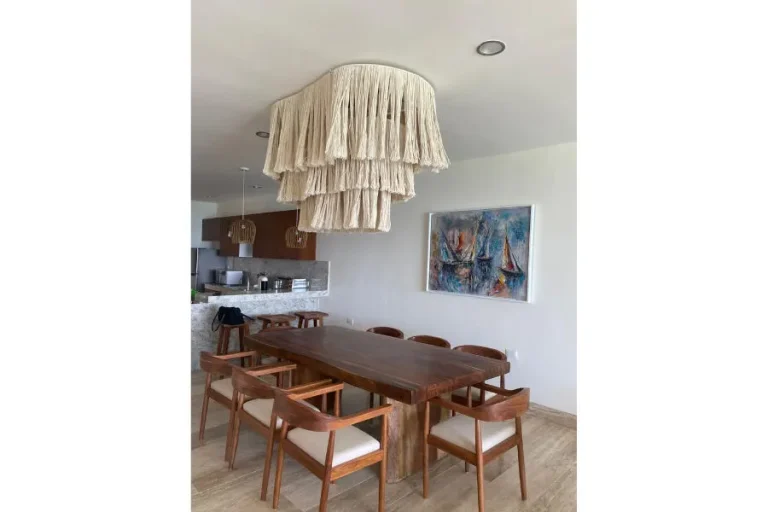 Mesa de comedor de madera con sillas de cojines claros, lámpara de flecos y cuadro junto a la cocina abierta.