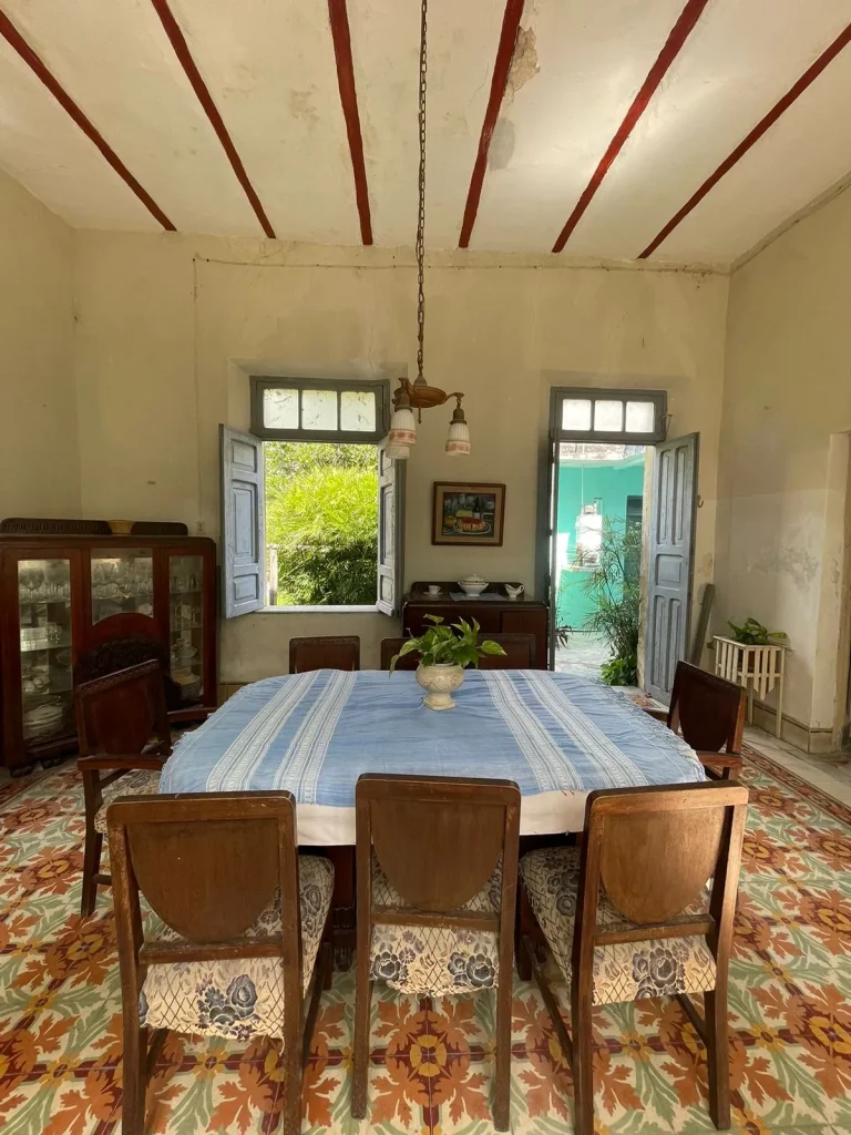 Comedor con sillas de madera, mantel azul, piso de mosaico floral y ventanas abiertas con contraventanas azules.