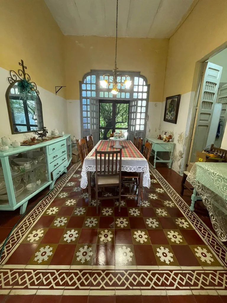 Comedor con mesa rectangular, sillas de madera, aparador verde claro y piso de losetas con motivos florales.