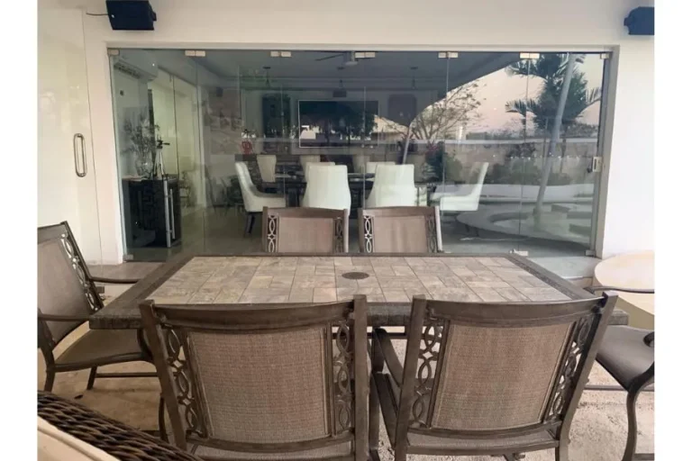 Mesa rectangular con sillas de exterior en una terraza, frente a puertas de cristal que permiten ver una barra interior.