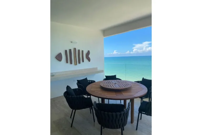Mesa redonda de madera con sillas oscuras en una terraza cubierta con barandal de cristal y vista al mar.