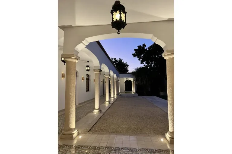 Corredor con arcos blancos, columnas de piedra y faroles encendidos junto a un patio al anochecer.