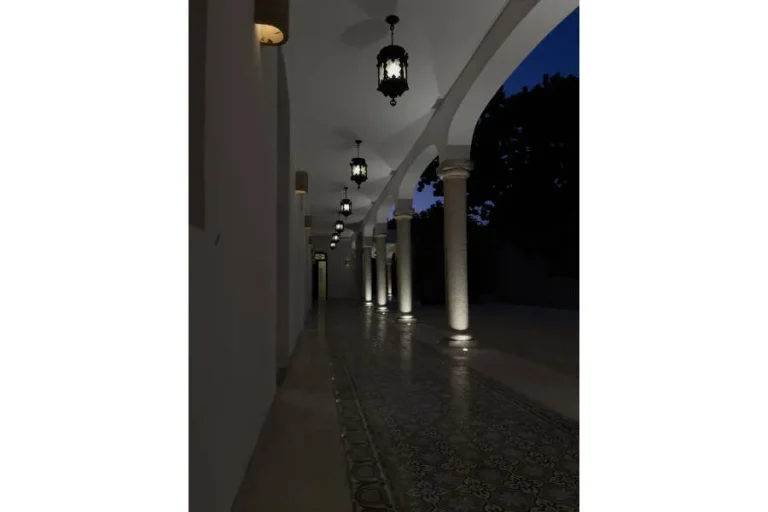 Corredor nocturno con arcos, columnas iluminadas, faroles colgantes y piso de mosaicos decorados.