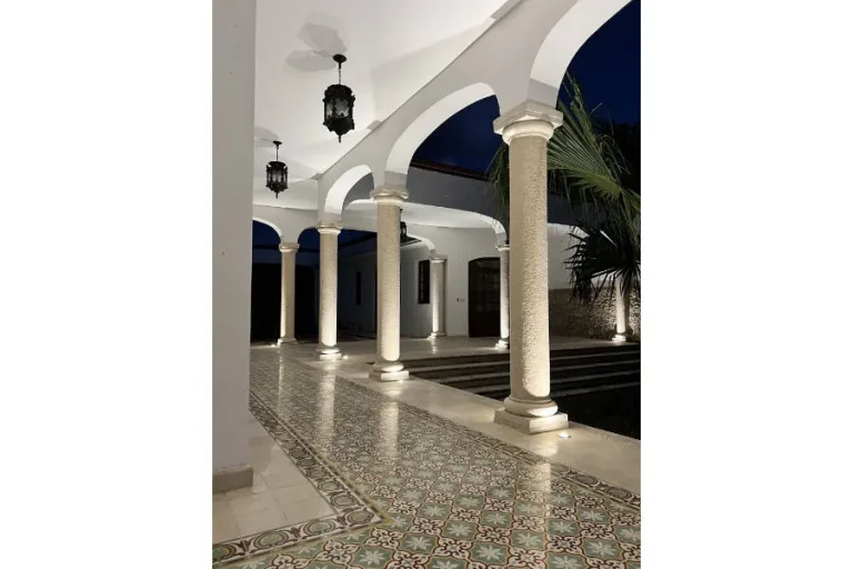 Corredor iluminado de noche con arcos blancos, columnas de piedra, faroles y un piso de mosaicos decorados.