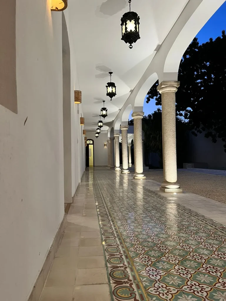 Corredor con arcos blancos, columnas iluminadas, faroles colgantes y piso de mosaicos con motivos florales.