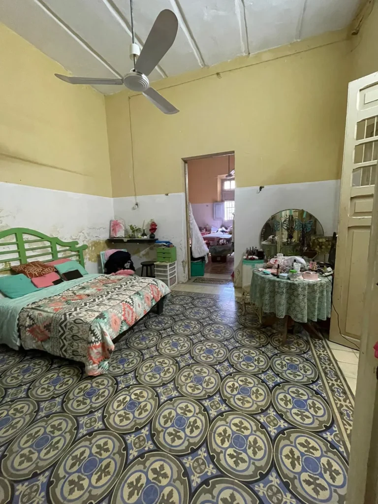Dormitorio con cama de cabecera verde, piso de mosaicos decorados, tocador con espejo y ventilador de techo.