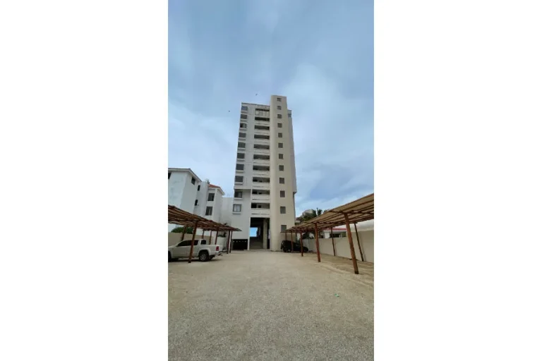 Edificio alto de fachada clara con balcones, frente a un patio de grava con espacios de estacionamiento techados.