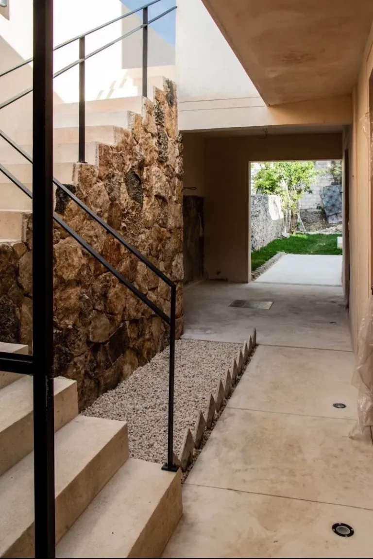 Escalera de tonos claros con barandal negro junto a un muro de piedra y un pasillo que conduce a un patio con césped.