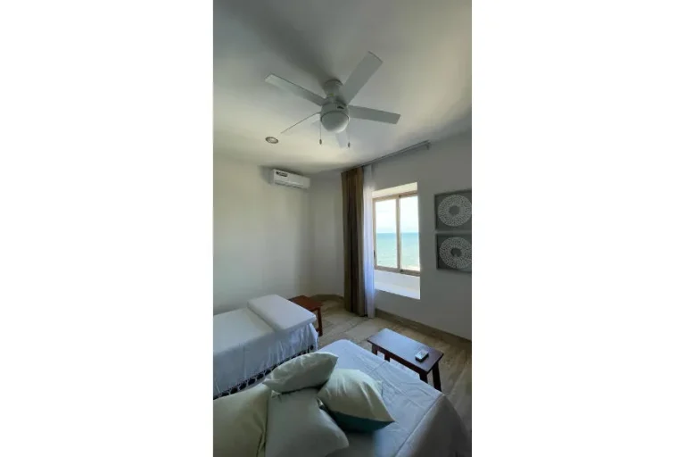 Estancia con sillones claros, cojines, mesa de centro, ventilador de techo y una ventana con vista al mar.