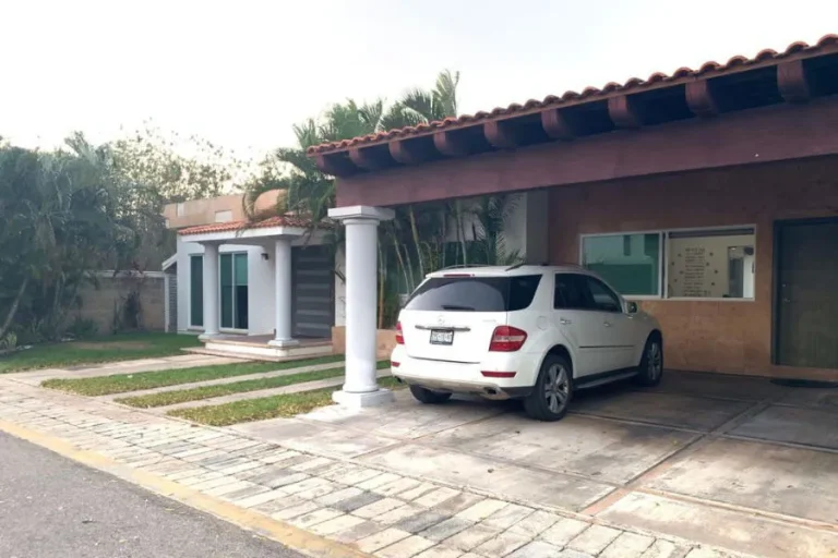 Fachada de vivienda con techo de tejas, columnas blancas, un vehículo blanco estacionado y franjas de césped en el acceso.