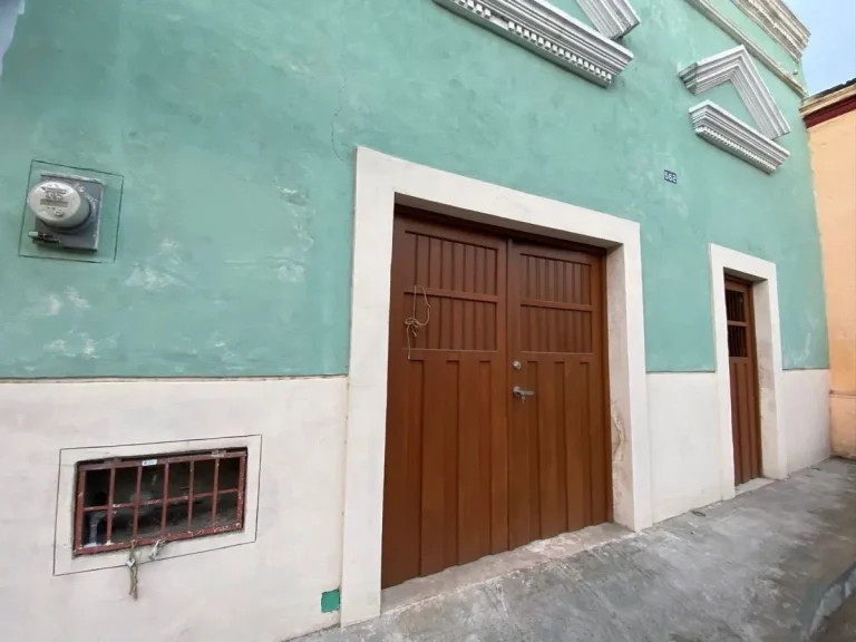 Fachada verde con zócalo blanco, portón y puerta de madera, ventana baja con reja y molduras en la parte superior.