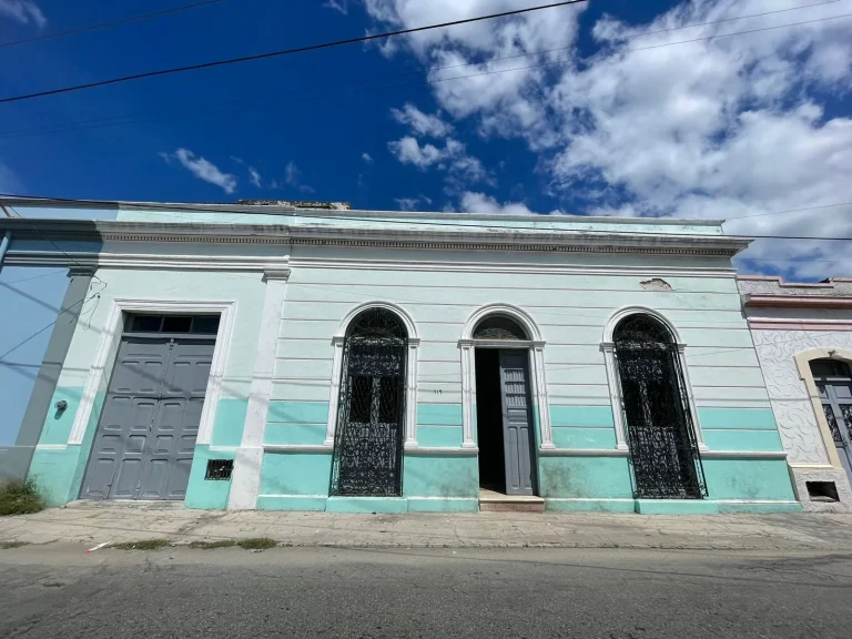 Fachada verde claro y turquesa con portón gris, puerta central y ventanas arqueadas con herrería negra.