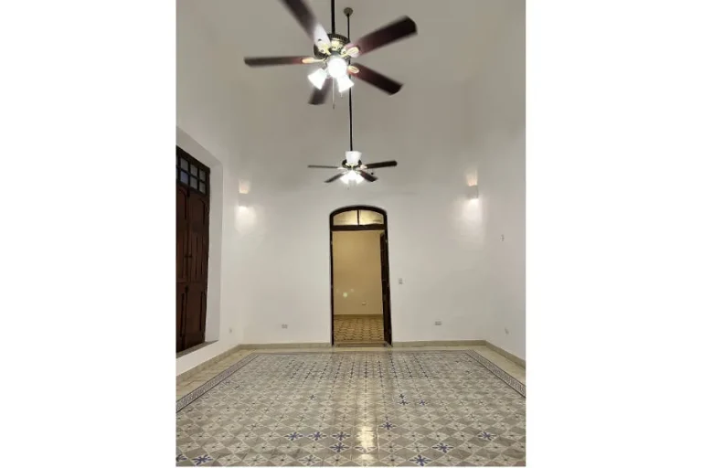 Habitación vacía con techo alto, dos ventiladores, piso de mosaicos y una puerta con remate en arco.
