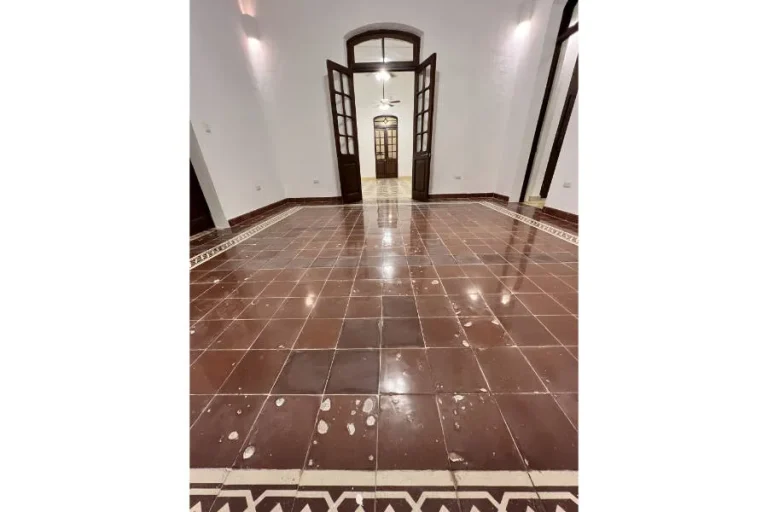 Habitaciones conectadas por puertas de madera abiertas, con paredes blancas y piso rojizo con manchas claras.