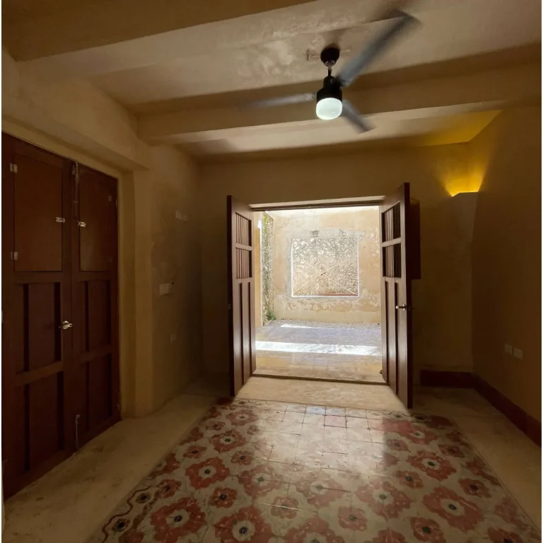 Interior de muros ocres con piso de motivos florales, ventilador de techo y puertas de madera abiertas a un patio.