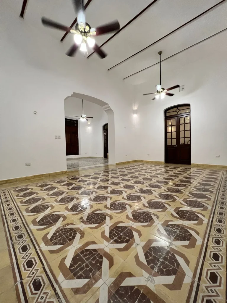 Interior vacío con piso de mosaico geométrico, techos altos, ventiladores, un arco entre ambientes y puertas de madera.