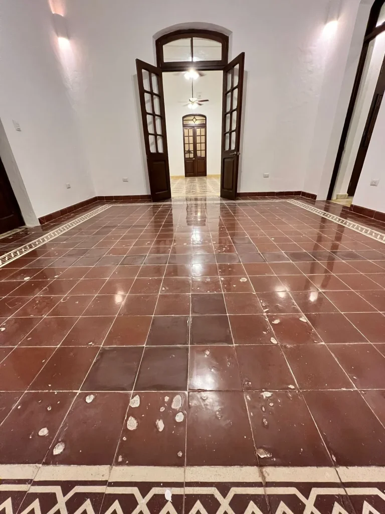 Habitación vacía con piso de losetas rojas desgastadas, cenefa geométrica y puertas de madera abiertas.