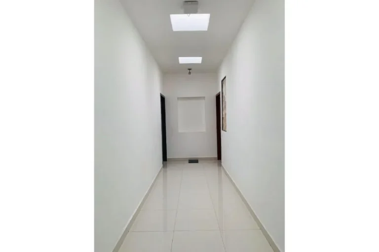 Pasillo de paredes blancas con piso cerámico brillante, luces en el techo y puertas laterales.