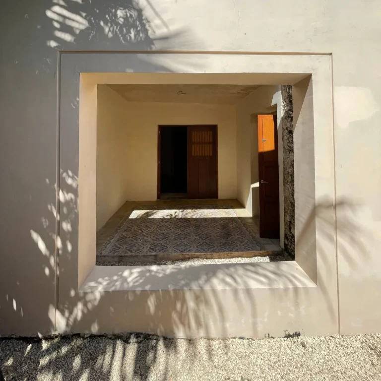 Abertura rectangular en un muro claro que enmarca un patio con piso decorativo, puertas de madera y un árbol.
