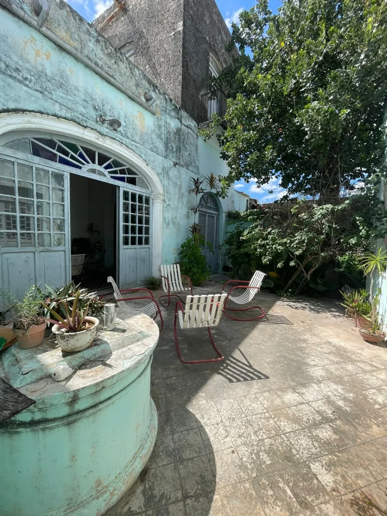 Patio con fachada verde claro, puertas de arco, sillas de jardín y macetas bajo la sombra de un árbol.