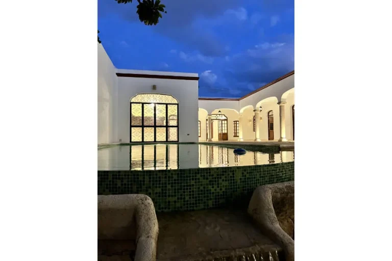 Piscina con borde de mosaico verde en un patio de muros blancos, arcos iluminados y cielo azul al atardecer.