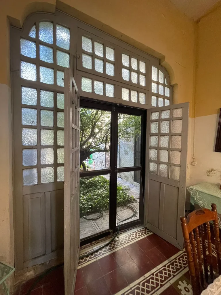 Puerta alta de madera con cristales texturizados, hojas abiertas y vista a un jardín arbolado.
