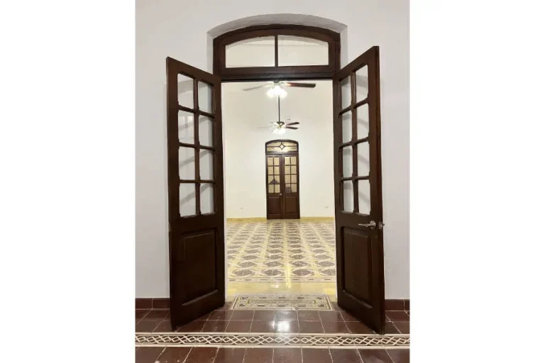 Puertas dobles de madera abiertas hacia una habitación con piso de mosaico, ventiladores de techo y otra puerta al fondo.