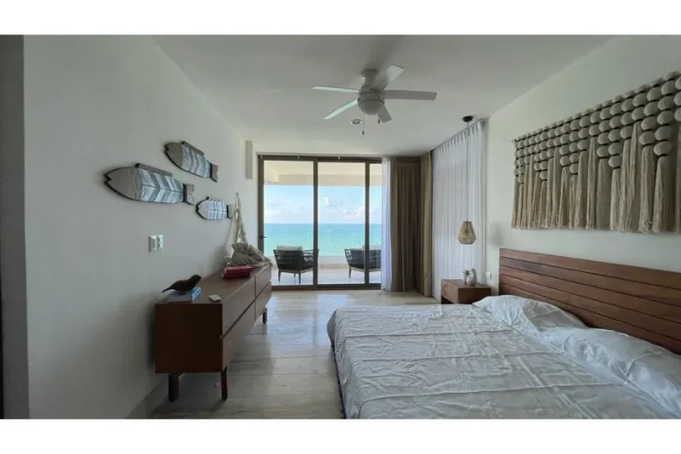 Recámara con cama doble, cabecera de madera y puertas de cristal hacia un balcón con vista al mar.