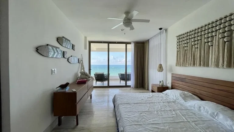 Recámara con cama doble, cabecera de madera, decoración de macramé y puertas de cristal hacia un balcón con vista al mar.