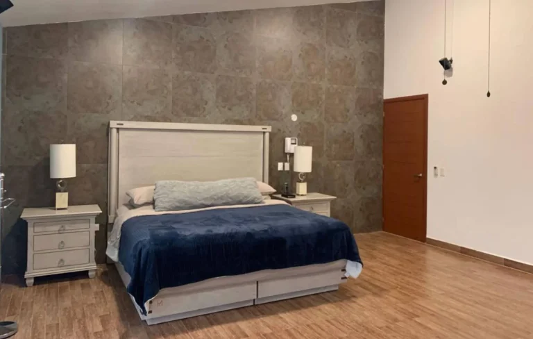 Recámara con cama de colcha azul, cabecera clara, dos burós con lámparas y muro de acabado pétreo.